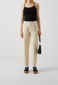 Débardeur noir ajusté, pantalon crème fuselé avec coutures verticales, baskets noires avec des rayures blanches, et un petit sac à main noir.