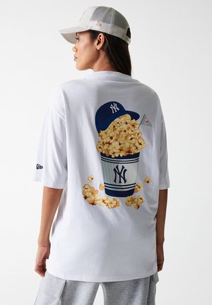 T-shirt surdimensionné blanc avec un graphisme d'un seau de pop-corn et d'une casquette bleue, avec du pop-corn jaune qui déborde. Manches courtes et coupe décontractée.