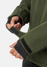 Grüne Jacke mit einem Klettverschluss am Ärmel, schwarzer innerer Fütterung und verstellbarem Ärmelausschnitt mit roten Nähten.