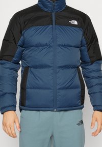 Padded jacket with a blue body and black shoulders, featuring a high collar, zip closure, and logo on the left chest. Textured fabric.