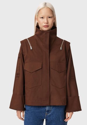 Osoba z długimi blond włosami w brązowej kurtce z wysokim kołnierzem, kieszeniami z patkami, detalami zamka błyskawicznego oraz w niebieskich dżinsach na jednolitym tle.