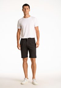 Schwarze Shorts mit umgeschlagenem Saum, Knopfverschluss und zwei Seitentaschen, kombiniert mit einem schlichten weißen T-Shirt mit kurzen Ärmeln und weißen Sneakers.