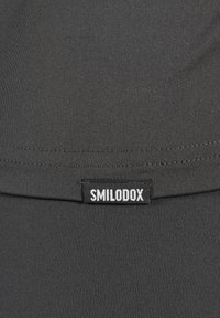 Gros plan sur un tissu noir avec une couture cousue et une petite étiquette noire rectangulaire portant la mention "SMILODOX" en lettres majuscules blanches.