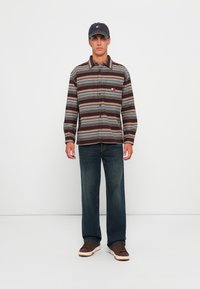 Bruine en blauwe gestreepte overhemd met knopen en een logo op de borst, gecombineerd met donkere jeans en bruine sneakers met witte zolen. Man staat tegen een effen achtergrond.