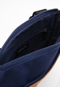 Wnętrze torby w kolorze brązowym i granatowym z wieloma kieszeniami na suwak, gładką tkaniną i widoczną metką Armani Jeans.