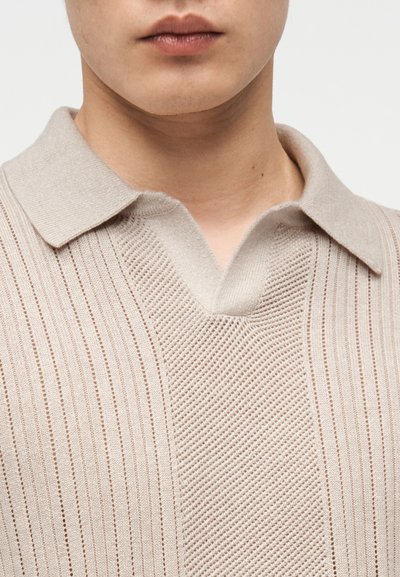 Κοντινό πλάνο ενός ατόμου που φοράει πλεκτό μπεζ πόλο πουκάμισο με υφή κατακόρυφης ρίγας και γιακά.