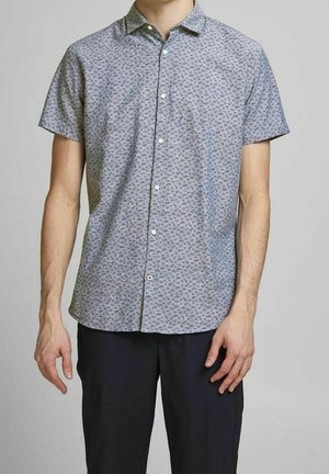 Homme portant une chemise à manches courtes gris clair à petits motifs floraux et un pantalon foncé, debout devant un fond uni.