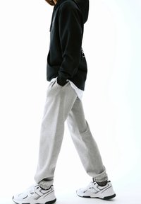 Zwarte hoodie, grijze sweatpants en witte sneakers. Hoodie heeft een logo; sweatpants hebben een losse pasvorm; het materiaal lijkt zacht.