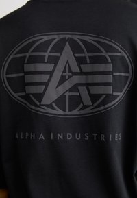 Tricou negru cu un logo mare, mat, care prezintă un design al globului și textul "ALPHA INDUSTRIES" într-o culoare contrastantă dedesubt.