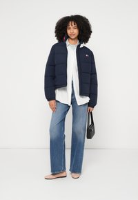 Jūras kara flotes pūdera jaka ar izšūtu dizainu, baltas krekls zemāk, gaiši zili džinsi un rozā dzīvokļi. Tur melnu somu.