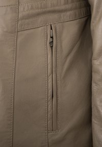 Beige Lederjacke mit glatter Textur, ausgestattet mit einer vertikalen Reißverschlusstasche und detaillierten Nähten entlang der Schulter.