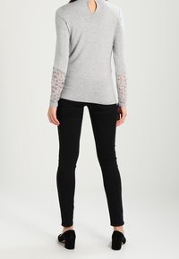 Grå långärmad topp med spetsaccenter på ärmarna, med en nyckelhålsöppning på baksidan, kombinerad med svarta åtsittande byxor och svarta klackar.