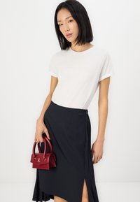 Tricou alb cu mânecă scurtă, fustă închisă la culoare cu un slit pe lateral și detalii cu logo, asortată cu o mică geantă burgundă. Fundal simplu.