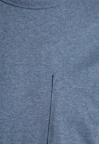Nærbillede af en blå heather-farvet T-shirt, der viser halsudskæringen og en lille brystlomme på højre side.