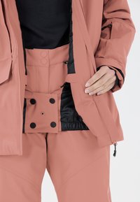 Vêtements d'extérieur résistants à l'eau avec une coque rose, une doublure intérieure noire et une taille à pression, dotés d'un tissu texturé et d'un design ergonomique.