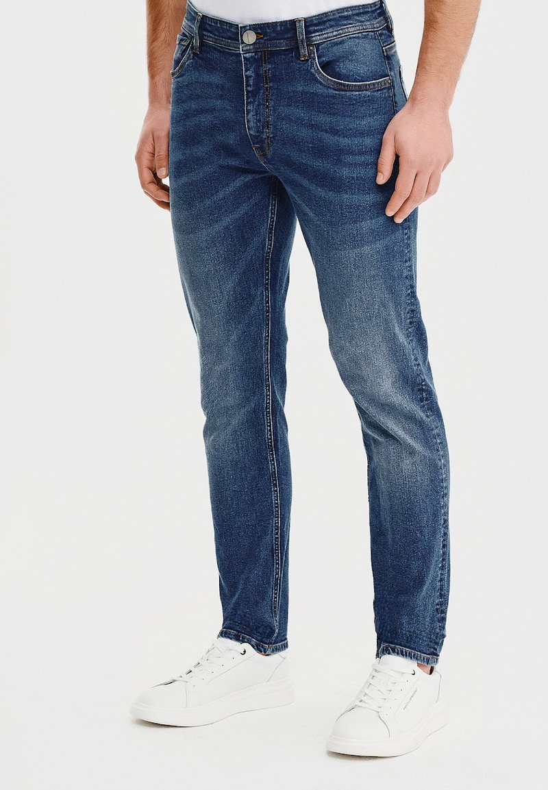 Ciemnoniebieskie dżinsy w slim fit, z delikatnym efektami przetarcia i lekkim blaknięciem. Stylizowane z białymi sneakersami i prostą białą koszulą.