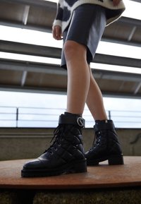 Botas de tobillo de cuero acolchado negro con suela gruesa y herrajes decorativos de plata. Presentan puntera reforzada y diseño de cordones.