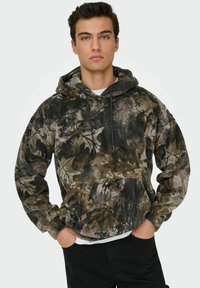 Hoodie camouflage en tissu doux et texturé. Comprend une poche avant, une capuche avec cordon de serrage, et un mélange de tons terreux et de motifs de feuilles.