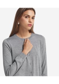 Kobieta z długimi brązowymi włosami, nosząca srebrne kolczyki w kształcie kół i szary kardigan zapinany na guziki, trzymająca górny guzik blisko swojej szyi.