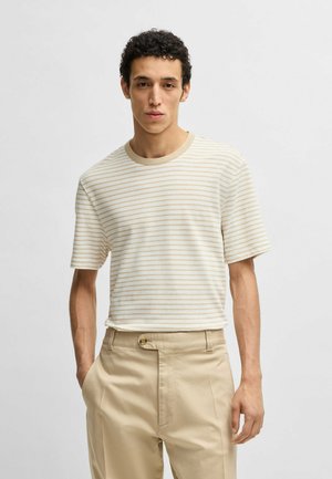 T-shirt a righe con maniche corte in crema e beige chiaro. Tessuto morbido, scollatura rotonda, vestibilità rilassata. Indossato con pantaloni beige chiaro con bottone.