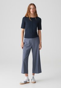 Navy blauw T-shirt met korte mouwen en gegolfde randen, gecombineerd met grijze wijde broek en witte sneakers met navy strepen. Soepele, casual uitstraling.