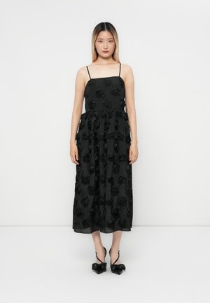 Black maxi dress featuring 3D rose patterns, spaghetti straps, and a fitted bodice. Paired with glossy high-heeled shoes.