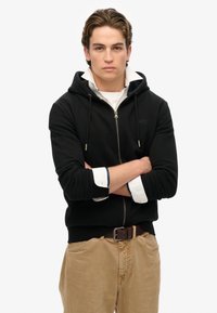 Schwarze Zip-Hoodie aus Baumwolle, mit verstellbarem Kapuzenzug und vorderen Taschen, getragen über einem weißen Hemd mit Kragen und beigen Hosen.