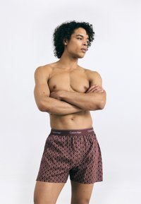 Sarkans rakstainais bokseru šorts ar Calvin Klein jostu. Audums ir mīksts un brīvi pieguļošs, ar ģeometrisku rakstu visā virsmā.