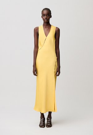 Mujer de pie con un vestido largo amarillo sin mangas con detalles de botones laterales y tacones abiertos con lazada negra sobre un fondo gris claro.