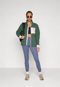 Vihreä fleecetakki, jossa vaaleanpunaiset yksityiskohdat, musta urheilurintaliivit, siniset legginsit ja beiget lenkkarit; malli pitelee mustaa reppua.