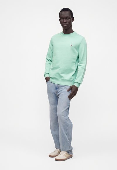 Homme portant un sweat-shirt vert menthe avec un petit logo zèbre, un jean bleu clair et des baskets beiges, debout avec une main dans la poche.