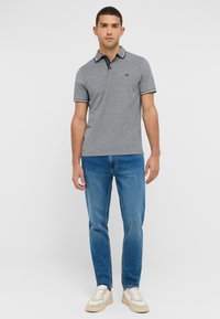 Pelēks polo krekls ar tumši zilu apkakli, īsām piedurknēm un baltām akcentiem, savienots ar zilajiem džinsiem un bēšajiem sporta apaviem. Vienkāršs, piegulošs dizains.