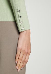 Świetlisty zielony mankiet swetra z prążkowanego materiału z czterema metalowymi guzikami, zestawiony z gładką, neutralną brązową tkaniną pod spodem.