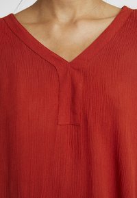 Haut rouge avec un col en V, tissu texturé aux motifs légèrement froissés et une petite patte centrale.