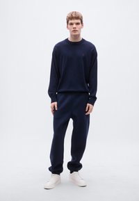 Μπλε ναυτικό πουλόβερ και τζόγκερς από μαλακό ύφασμα, με στρογγυλή λαιμόκοψη και ελαστικά μανίκια, συνδυασμένα με λευκά αθλητικά παπούτσια.