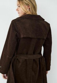 Brązowy sztruksowy płaszcz z szerokim kołnierzem, paskiem w talii oraz pionowym prążkowanym wzorem. Projekt zawiera mankiety zapinane na guziki oraz luźny krój.