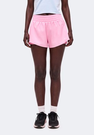 Personne portant un short de sport rose clair, des chaussettes blanches, des chaussures de course noires et roses, et un haut court blanc, debout devant un fond uni.