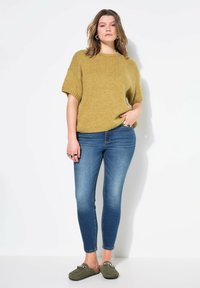 Hořčicový pletený svetr s krátkými rukávy, kombinovaný se modrými skinny džíny a olivově zelenými botami na slip s dekorativním kováním.