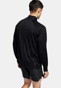 Haut de sport noir à manches longues avec un col haut, en tissu texturé et accents réfléchissants subtils sur les manches et le logo.