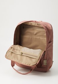 Sac à dos en tissu rose avec un intérieur beige, comportant des poches zippées et des compartiments d'organisation. L'extérieur a une texture lisse.