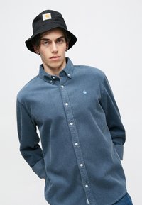 Modrá kordurová košeľa s gombíkmi a klasickým golierom, na ktorej sa nachádza malá svetlomodrá značka a biele gombíky. Čierny klobúk typu bucket so značkou.