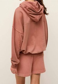 Sweat à capuche rose poussiéreux surdimensionné avec capuche réglable, poignets côtelés et short assorti. Confectionné en tissu doux et texturé avec des coutures apparentes.