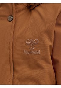 Die braune Stoffjacke hat eine glatte Textur und ist mit einem gestickten Logo in hellerem Farbton versehen. Sie hat einen Metallknopf mit einem dekorativen Design.