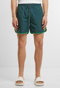 Férfi téglalap és homokszínű sportnadrágban, zöld díszítéssel és fehér slip-on szandálban áll, egy sima fehér háttér előtt.