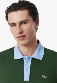 Pikétröja med grön kropp, ljusblå krage och tre knappar samt en liten broderad logotyp på vänster bröst. Strukturerat tyg.