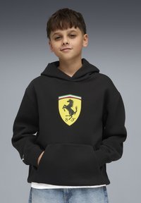 Sweat à capuche noir en mélange de coton, arborant un logo Ferrari jaune avec un cheval cabré. Comprend une poche avant et des poignets côtelés.