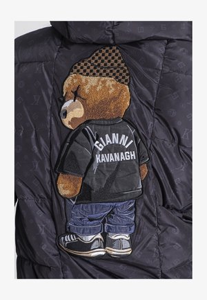 Chaqueta negra acolchada con un parche texturizado de un oso que lleva una camiseta negra con "GIANNI KAVANAGH" en la espalda y pantalones de mezclilla azules.