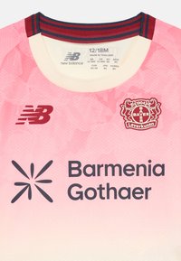 Camisola de futebol rosa com decote redondo, apresentando um design em degradê, logotipo da New Balance e emblema do Bayer Leverkusen. O texto inclui "Barmenia Gothaer."