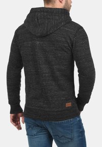 Un sweat à capuche gris foncé en tissu texturé, doté d'une capuche et d'un petit patch logo dans le bas du dos. Associé à un jean bleu.