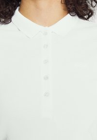 Valkoinen polo paita tekstuurilla, jossa on klassinen kaulus, kuusi läpinäkyvää nappia ja huomaamaton logo kirjailtuna rintaan.